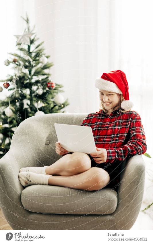 Glückliches Teenager-Mädchen mit Weihnachtsmannmütze und Laptop in einem weihnachtlich dekorierten Zimmer, das sich mit Freunden und Verwandten unterhält
