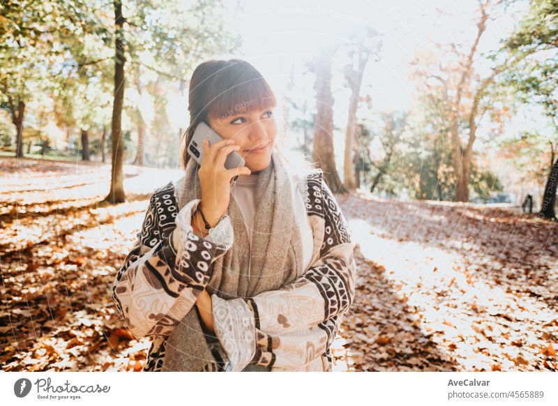 Happy girl using smart phone during Autumn in a park taking selfie, woman talking on mobile in fall. Videoanruf im Freien, gute Nachrichten erhalten. Person