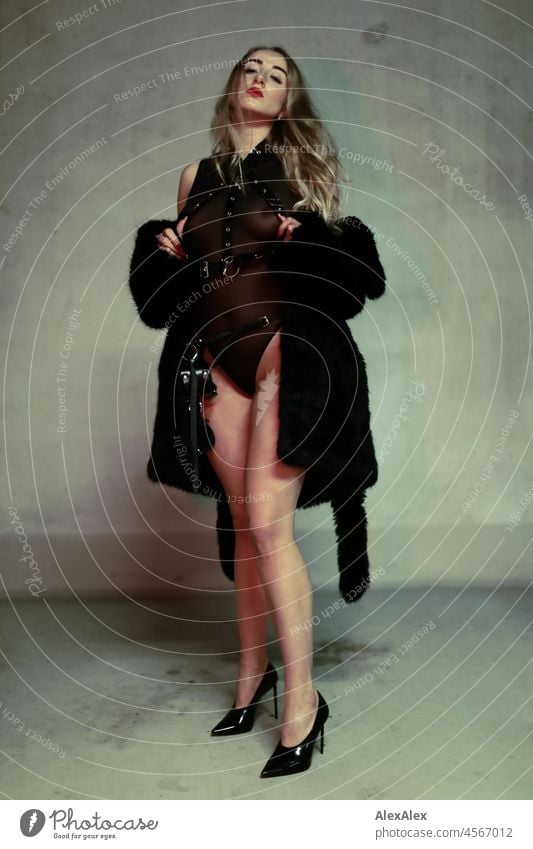 Portrait einer jungen, blonden Domina in High Heels vor einer Betonwand. Sie trägt schwarze Unterwäsche und und einen schwarzen, kurzen Mantel Frau junge Frau
