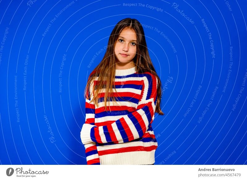 Adorable elf Jahre altes Mädchen trägt einen roten und blauen gestreiften Pullover auf blauen Studio Hintergrund, hübsche braunhaarige Pony Frisur europäisches Aussehen Kind Pose Innen lächelnden Blick in die Kamera, Generation Z Konzept