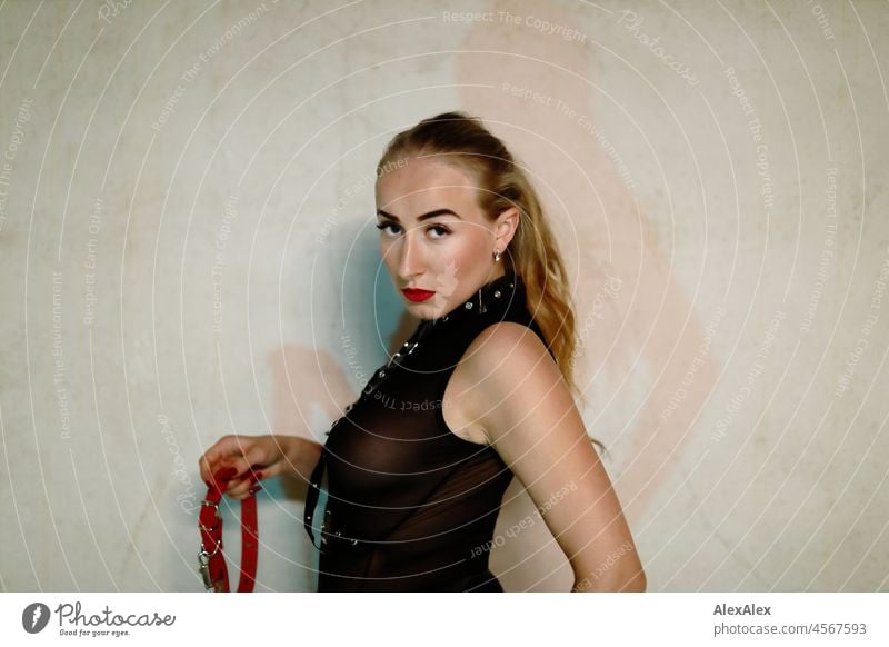 Portrait einer jungen, blonden Domina vor einer Betonwand. Sie trägt schwarze Unterwäsche und hält ein rotes Lederhalsband auffordernd in der Hand und schaut sehr ernst in die Kamera