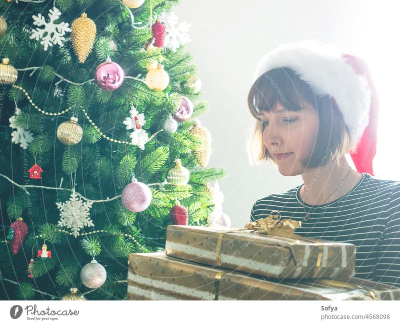 Frau mit Weihnachtsmannmütze hält eingepacktes Geschenk, dahinter Weihnachtsbaum Weihnachten Geschenke Baum Morgen auspacken Überraschung Feiertag aufgeregt
