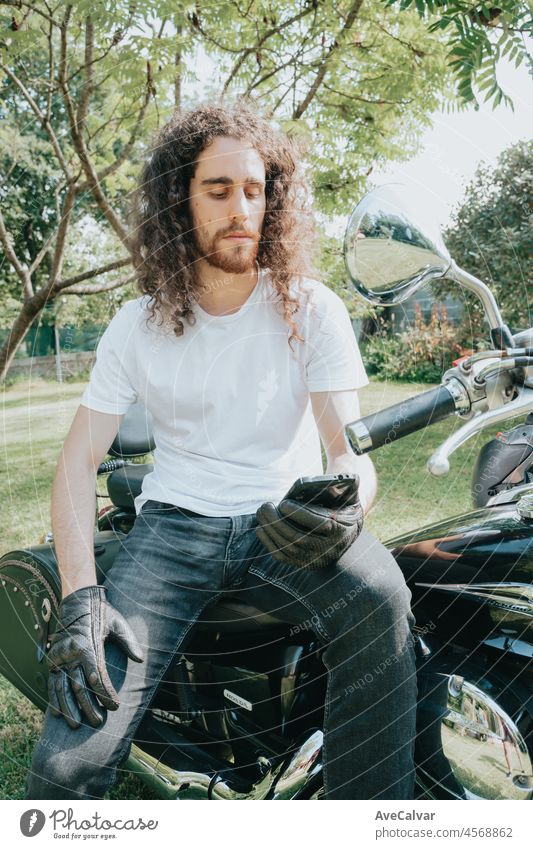 Junge lange Haare Motorrad Kerl überprüft sein Telefon, während auf seiner alten Schule Motorrad während einer Pause von der Straße Route sitzen. Liberty Leben, junger Mann Heavy Metal, weißes T-Shirt und Handschuhe.