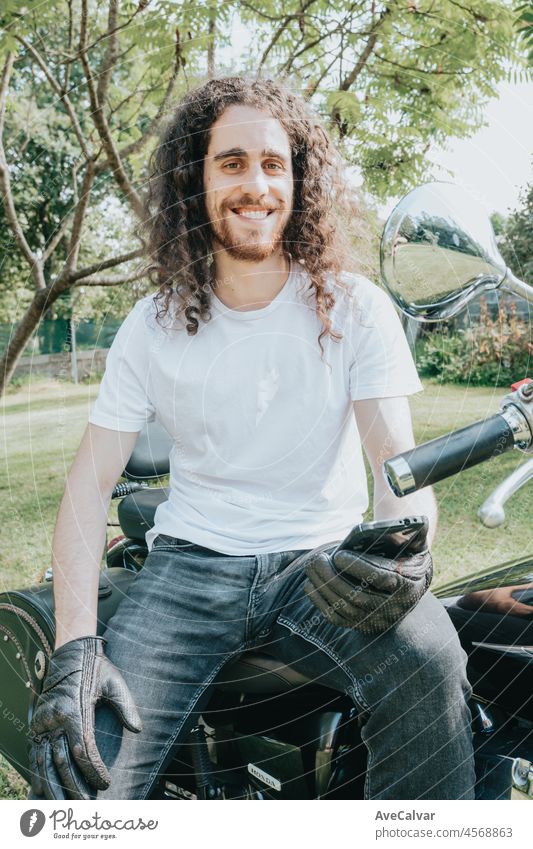 Junge lange Haare Motorrad Kerl lächelnd in die Kamera, während sein Telefon zu überprüfen, während auf seiner alten Schule Motorrad während einer Pause von der Straße Route sitzen. Liberty Leben, junger Mann Heavy Metal, weißes T-Shirt
