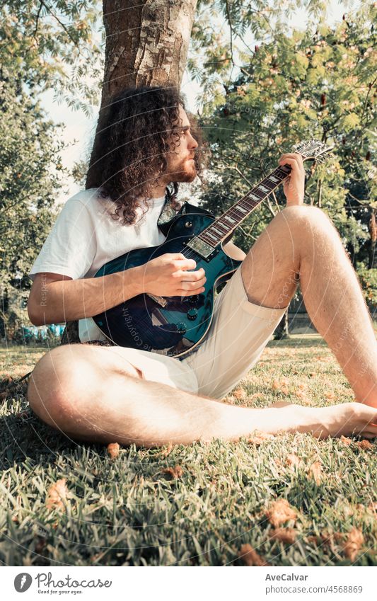 Junge lange Haare Hippie-Mann lächelnd Gitarre spielen im Park, während gegen einen Baum ruht. Hobby-Kunst-Aktivität für junge Erwachsene. Authentische Männlichkeit Moment.