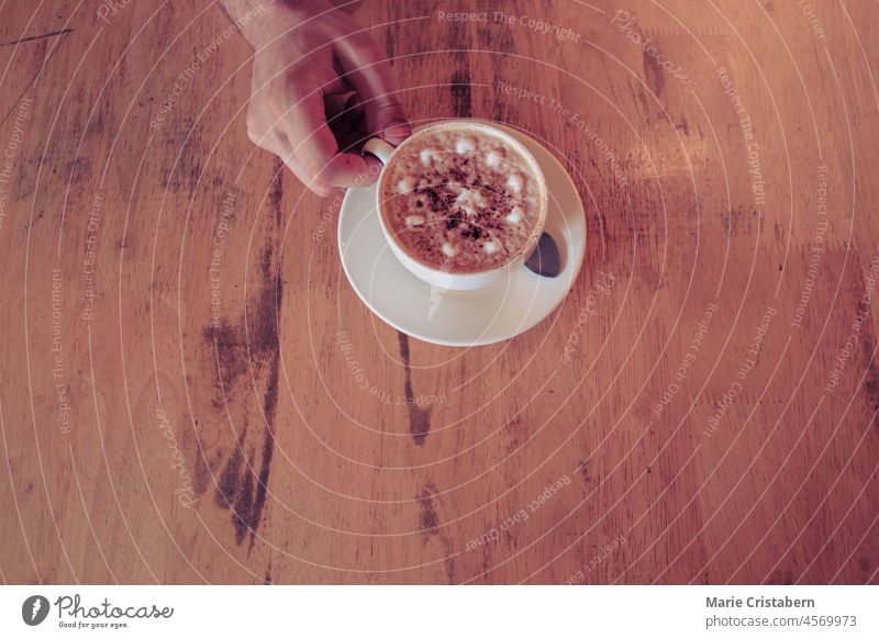 Draufsicht auf eine Hand, die eine frisch zubereitete Tasse mit schaumigem Cappuccino in einem Café hält Kaffeehauskultur Textfreiraum Hintergrund Getränk