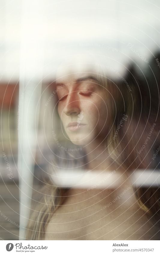 Aktportrait einer jungen, blonden, langhaarigen Frau, die nackt und mit geschlossenen Augen hinter der Fensterscheibe steht Jugendlichkeit gesund