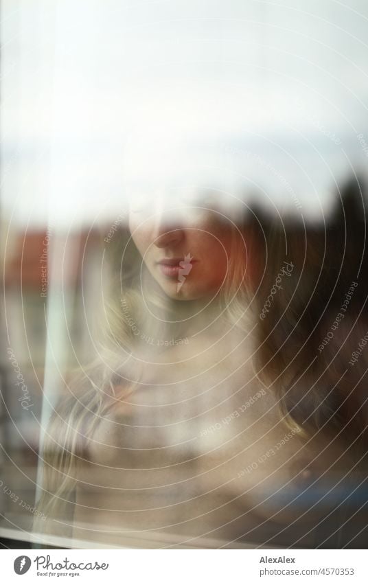 Aktportrait einer jungen, blonden, langhaarigen Frau, die hinter der Fensterscheibe nackt nach unten schaut Jugendlichkeit gesund Gesichtsausdruck Wärme drinnen