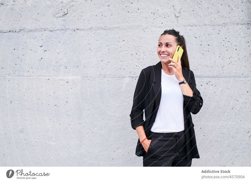 Lächelnde Frau, die mit ihrem Smartphone an der Wand spricht heiter sprechen Funktelefon reden Telefonanruf Gespräch benutzend Straße Apparatur formal