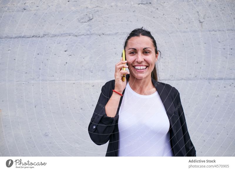 Lächelnde Frau, die mit ihrem Smartphone an der Wand spricht heiter sprechen Funktelefon reden Telefonanruf Gespräch benutzend Straße Apparatur formal Arbeiter