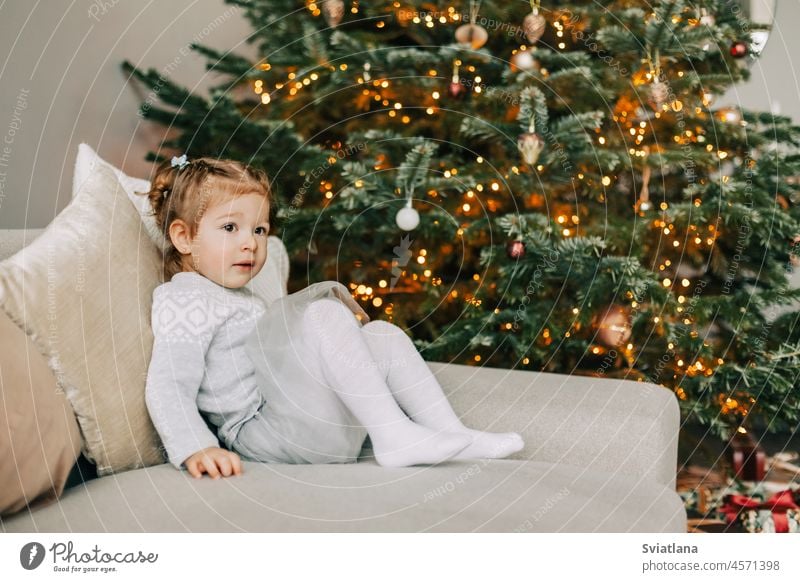 Ein hübsches Mädchen mit einem Hut sitzt auf dem Sofa in der Nähe eines Weihnachtsbaums, gekleidet für den Urlaub, das Kind lächelt und freut sich über das Geschenk