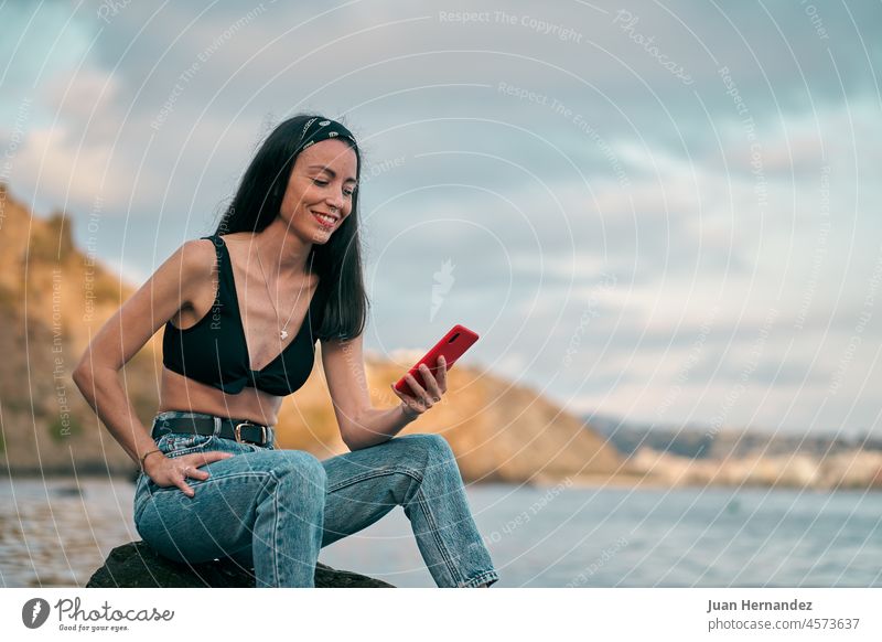 Junge Frau schaut auf ihr Smartphone und sitzt auf einem Felsen am Strand hübsch Blick Sitzen jung Hintergrund Handy eine Person stylisch Halt horizontal Mobile