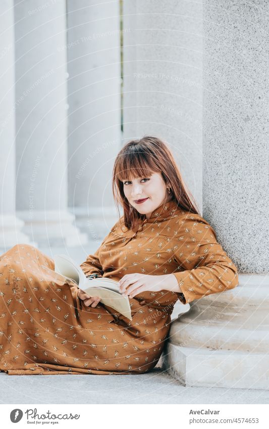 Junge Frau liest ein Buch und lächelt in die Kamera, während sie auf dem Boden eines architektonischen Ortes sitzt. Glücklich beim Lernen und Lesen von Abenteuern. Raum kopieren. 80er-Jahre-Stimmung. Trendy Styling Kleid
