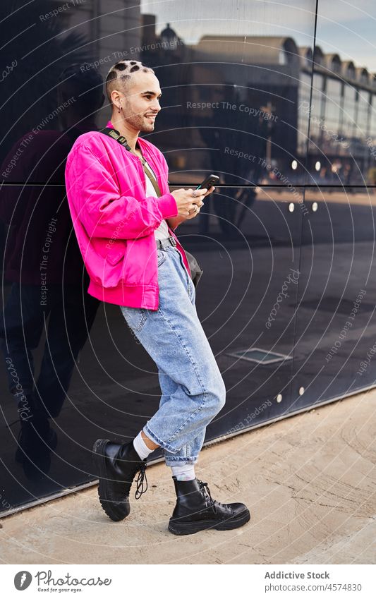 Stilvoller junger Mann, der in der Nähe einer reflektierenden Wand auf seinem Smartphone surft exzentrisch Straße benutzend cool Nachricht modern