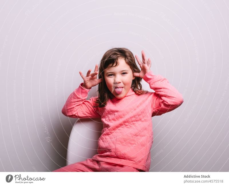 Lustiges kleines Mädchen mit Gesicht Grimasse aufgeilen Zunge zeigen lustig gestikulieren Glück Stuhl sitzen Kind Porträt heiter ausspannen Wand spielerisch