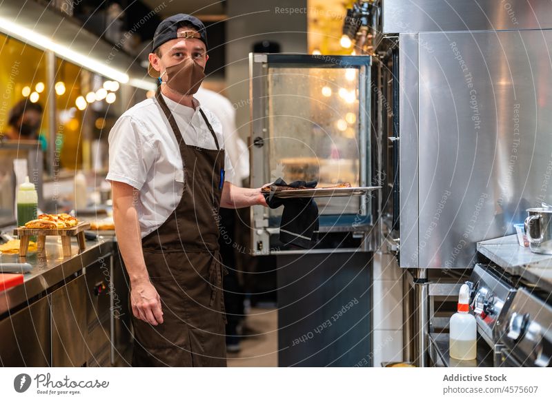 Koch, der den Teller mit dem Essen in den Ofen stellt Mann vorbereiten Lebensmittel backen Mundschutz Küche Vorrichtung COVID19 männlich Restaurant kulinarisch