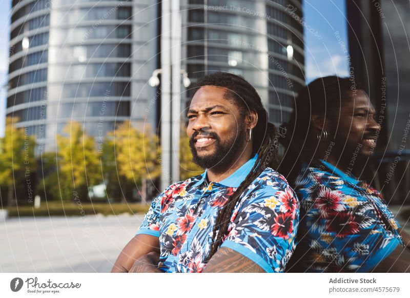 Schwarzer Mann sitzt auf der Straße in der Nähe eines Gebäudes urban Großstadt modern Stil Lächeln Zeitvertreib Bekleidung Glück Outfit Reflexion & Spiegelung
