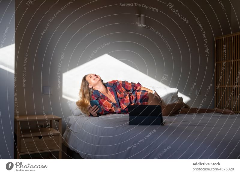 Fröhliche Frau auf dem Bett liegend mit Smartphone und Tablet Lachen Glück Tablette benutzend Funktelefon Kälte Schlafzimmer ruhen Browsen Komfort Apparatur