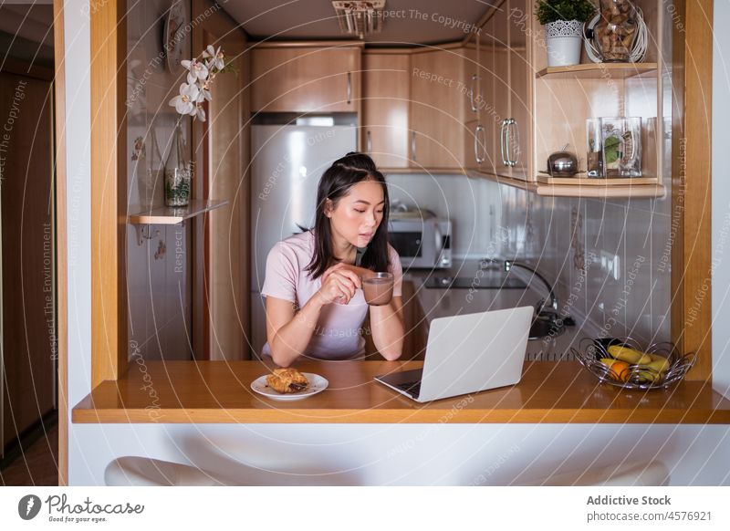 Asiatische Frau mit Getränk beim Surfen auf dem Laptop Abfertigungsschalter Küche Browsen online Frühstück Heißgetränk Internet Gebäck Lebensmittel Bäckerei