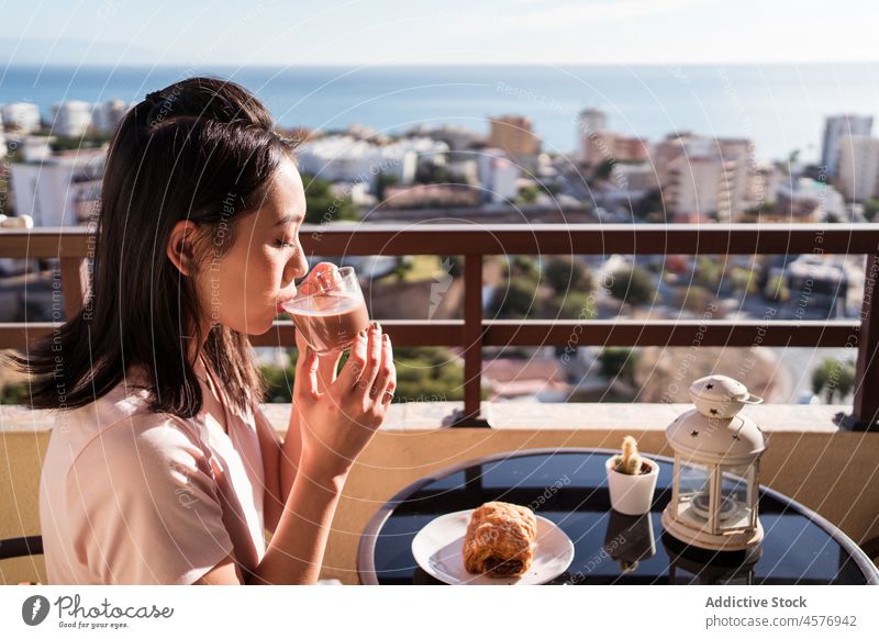 Fröhliche asiatische Frau mit Getränk auf dem Balkon trinken Frühstück Terrasse Morgen Heißgetränk Stadtbild Erfrischung Gebäude Zaun Tisch lecker geschmackvoll