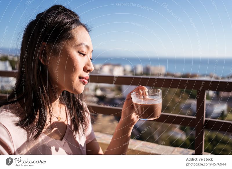 Fröhliche asiatische Frau mit Getränk auf dem Balkon trinken Frühstück Terrasse Morgen Heißgetränk Stadtbild Gebäude Zaun lecker geschmackvoll Großstadt