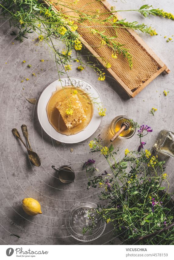 Ganze rohe Honigwabe mit Honig und Honiglöffel, frische Blumen und Zitrone auf grauem Betonküchentisch. Natürlicher gesunder Lebensstil. Ansicht von oben. ganz