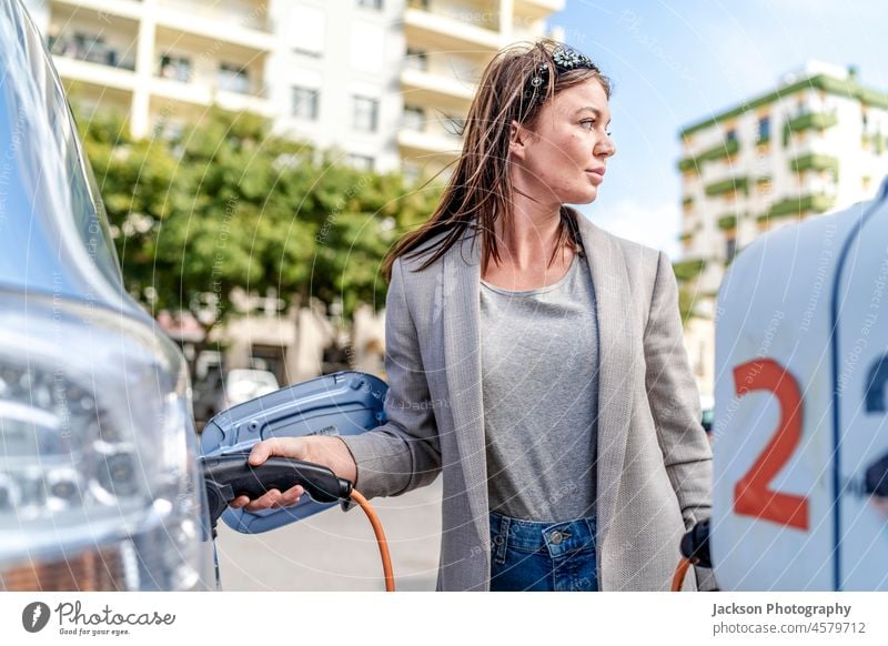Eine Frau lädt ein Elektroauto in einer städtischen Umgebung auf. PKW Gebühr Ladestation Kaukasier Erneuerbare Energie schön jung regenerativ Beteiligung