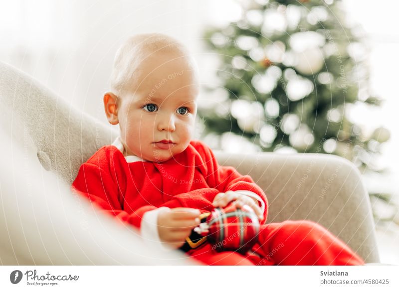 Ein als Weihnachtsmann verkleidetes Kind sitzt an Heiligabend auf einem Stuhl neben einem Weihnachtsbaum Weihnachten Baby Baum weiß Claus Weihnachtszeit