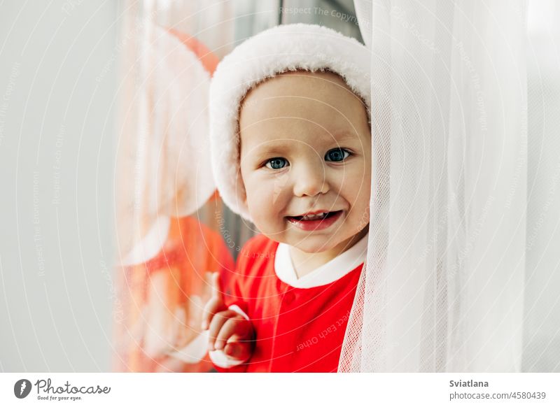 Porträt eines bezaubernden Babys in einem Weihnachtsmannkostüm am Fenster. Nahaufnahme. Das Konzept von Weihnachten und Neujahr Warten Hut Glück Fröhlichkeit