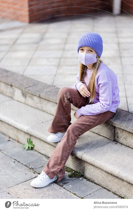Nette kleine kaukasische Mädchen acht Jahre alt mit blonden Haaren und schützende Gesichtsmaske zu Fuß im Freien. Kind trägt stilvolle Shirt und Strickmütze violette Farbe. Trendy Farbe des Jahres 2022 sehr peri