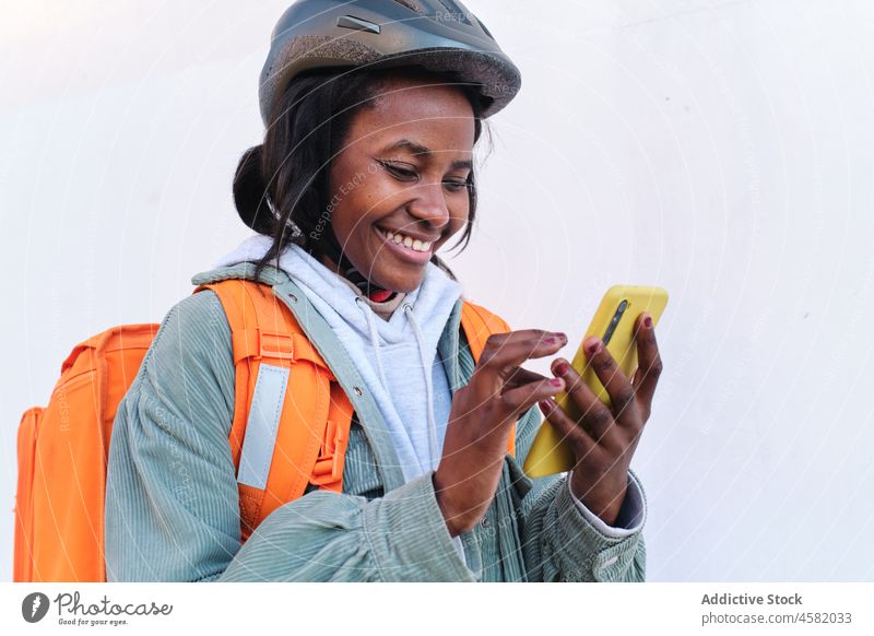 Glückliche schwarze Lieferfrau, die auf der Straße ihr Smartphone durchsucht Frau Versand Rucksack Dienst Orden Browsen prüfen Lächeln Arbeit Thermo Spaziergang