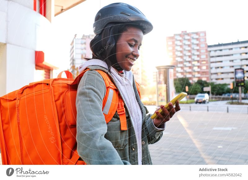 Glückliche schwarze Lieferfrau, die auf der Straße ihr Smartphone durchsucht Frau Versand Rucksack Dienst Orden Browsen prüfen Lächeln Arbeit Thermo Spaziergang