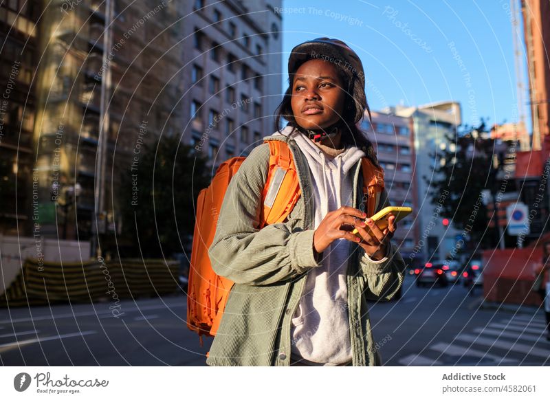 Schwarze Lieferfrau durchsucht Smartphone in der Nähe der Straße Frau Versand Rucksack Dienst App Orden prüfen Arbeit Fahrbahn Straßenrand Thermo Spaziergang