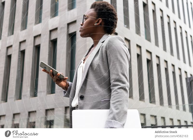 Schwarze Unternehmerin mit Laptop und Mobiltelefon auf der Straße in der Nähe eines Bürogebäudes Frau Gerät Smartphone Netbook Afroamerikaner Geschäftsfrau