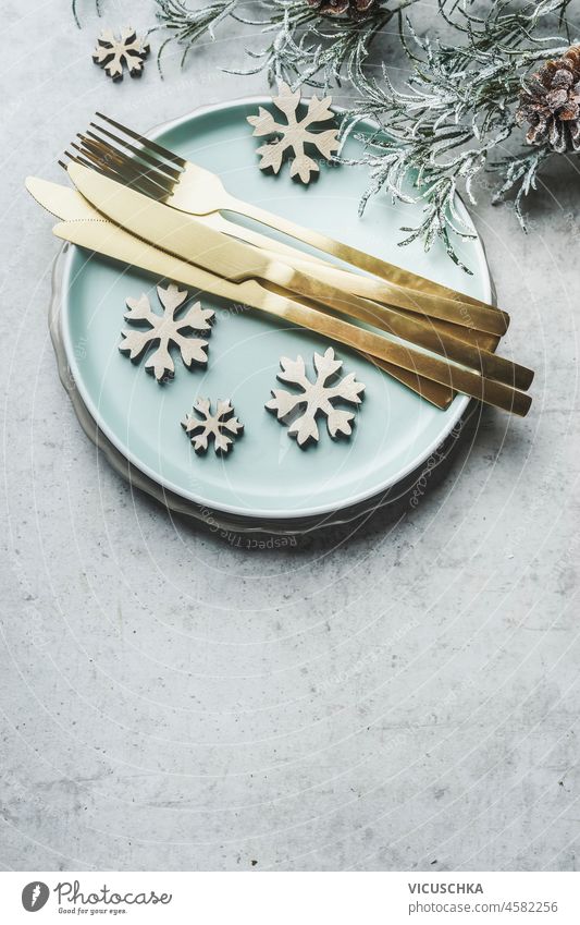 Weihnachtlich gedeckter Tisch mit hellblauem Teller, goldenem Besteck, Schneeflocken aus Holz, Tannengrün und Tannenzapfen auf grauem Betontisch. Festliche Tischdekoration für das Weihnachtsessen. Draufsicht mit Kopierbereich
