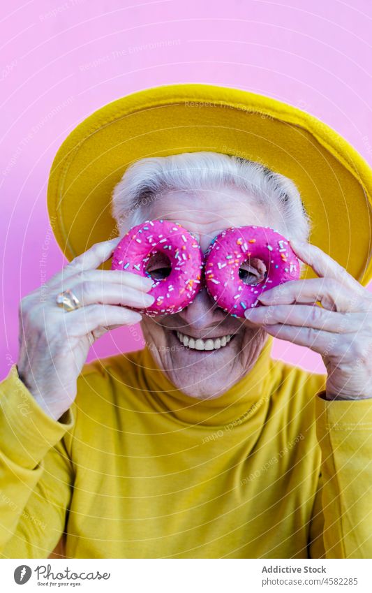 Ältere Frau bedeckt Augen mit Donuts Doughnut Augen abdecken Dessert Spaß haben sorgenfrei älter alt in den Ruhestand gehen Porträt süß Wand Stil gealtert Glück