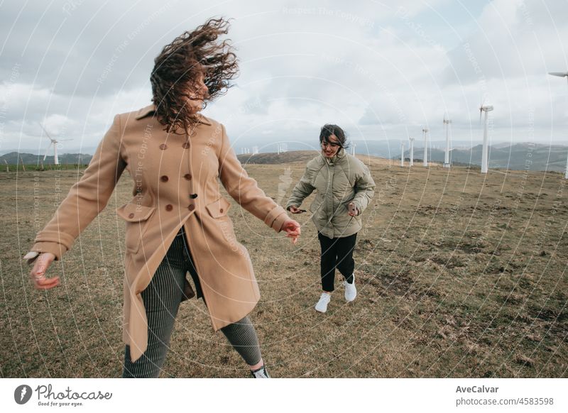 Zwei junge Frauen, in Winterkleidung an einem windigen Tag, genießen den Tag und haben Spaß beim Laufen zwischen elektrischen Windrädern. Lebensstil. Hipster und moderner Stil. Kino-Stil.