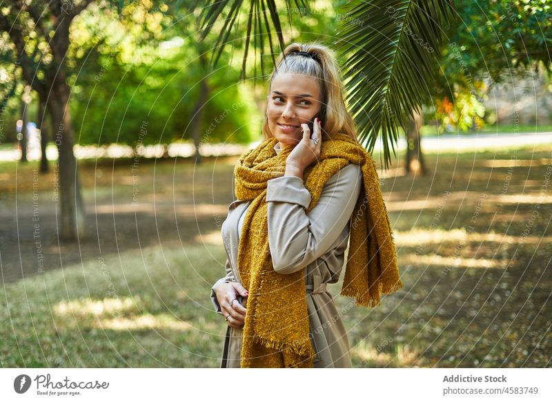 Lächelnde Frau mit warmem Schal, die im Park mit ihrem Handy telefoniert Smartphone reden sprechen Telefon Mobile Mantel Gespräch positiv Oberbekleidung blond