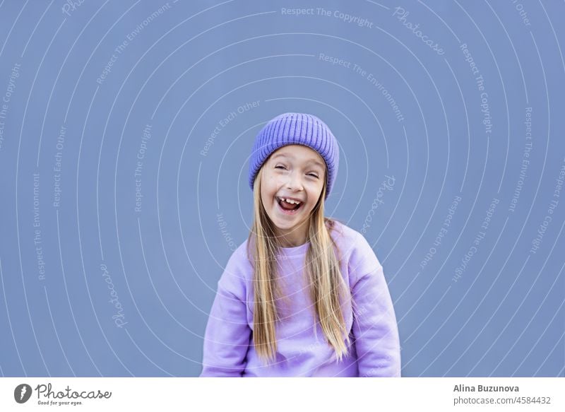 Nette kleine kaukasische Mädchen acht Jahre alt mit blonden Haaren lächelnd im Freien. Kind trägt stilvolle Shirt und Strickmütze violette Farbe. Trendy Farbe des Jahres 2022 sehr peri