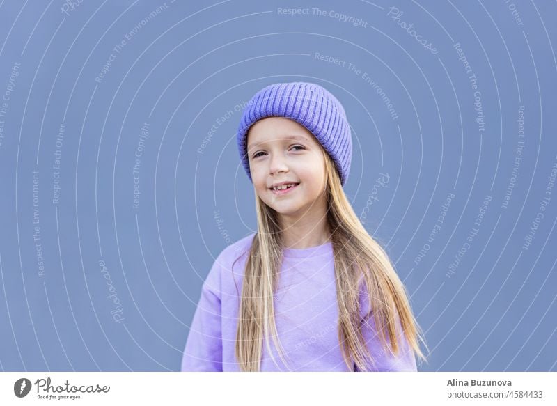 Nette kleine kaukasische Mädchen acht Jahre alt mit blonden Haaren lächelnd im Freien. Kind trägt stilvolle Shirt und Strickmütze violette Farbe. Trendy Farbe des Jahres 2022 sehr peri