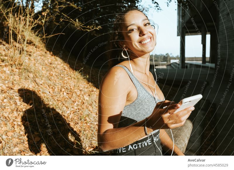 Lächelnd fit afrikanischen arabischen Frau in Tank-Top und Leggins nutzt sein Telefon während der Ruhe sitzen im Freien während eines Sonnenuntergangs, im Freien Stadion gehen, um Yoga zu üben, um nach stressigen Tag zu entspannen, mit Kopie Raum