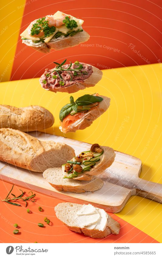 Kleine schwebende Baguette-Sandwiches auf leuchtend gelbem und orangefarbenem Hintergrund, verziert mit Grünpflanzen Belegtes Brot pulsierend grüne Blätter