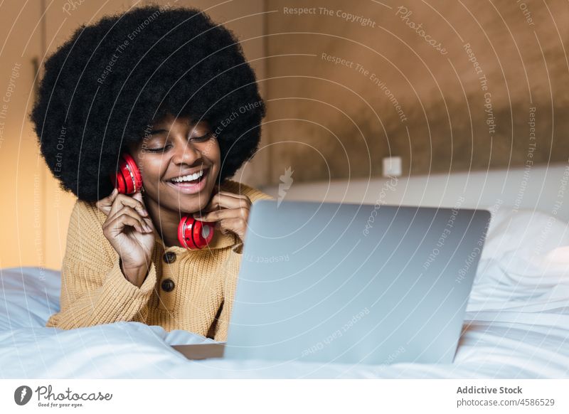 Lächelnde schwarze Frau, die mit einem Laptop im Bett Musik hört Ohrstöpsel benutzend heimwärts sich[Akk] entspannen ruhen Wochenende zuhören Afroamerikaner