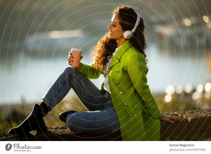 Hübsche junge Frau hört Musik mit Smartphone und Kaffee zum Mitnehmen am Fluss an einem sonnigen Herbsttag hübsch attraktiv Kaukasier im Freien schön