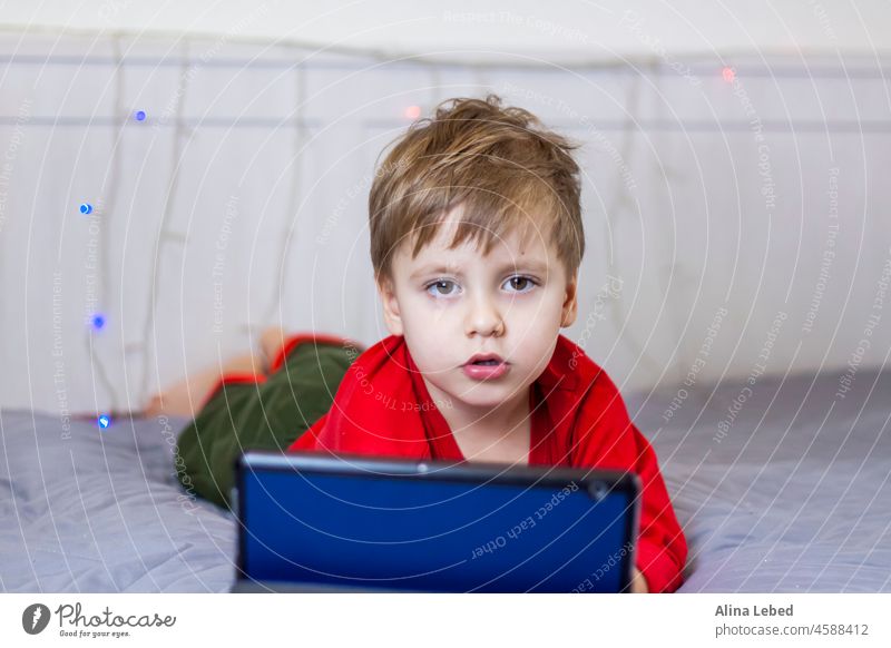 Ein süßer Junge liegt auf dem Bett und schaut auf ein Tablet. Online-Training, Kommunikation über das Internet. Lustiges lächelndes Kind. Schule Menschen