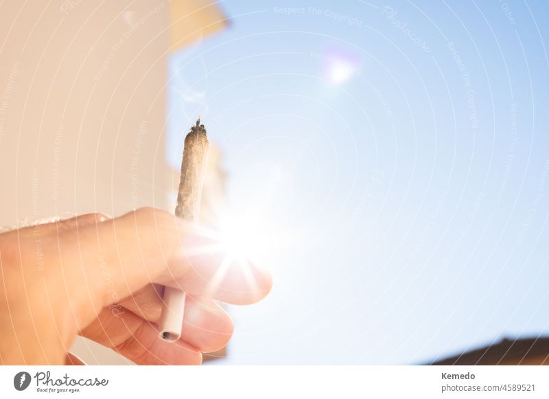 Hand mit einem Marihuana-Joint, im Hintergrund der Himmel und die Sonne. Gelenk Cannabis Unkraut Zigarette Textfreiraum sonnig im Freien Transparente horizontal