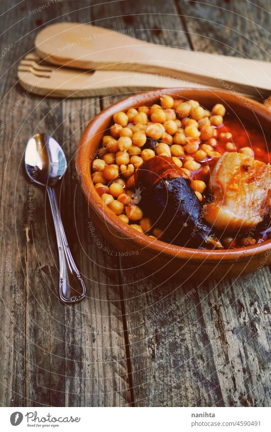 Kichererbsen, Wurst und Speck in einem Kochtopf mit Holzlöffel und Gabel und einem kleinen Metalllöffel. Typisches Essen aus Madrid, Spanien, mit einer rustikalen Holztafel als Hintergrund.