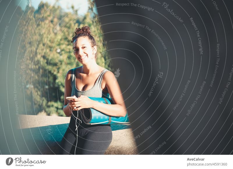 Lächelnde junge afrikanische Frau, die ihr Smartphone während des Trainings hält und benutzt, während sie draußen sitzt und eine Yogamatte hält, Musik hört und im Freien plaudert. Neue Gewohnheiten Lebensstil. Urban Sport Sonnenuntergang