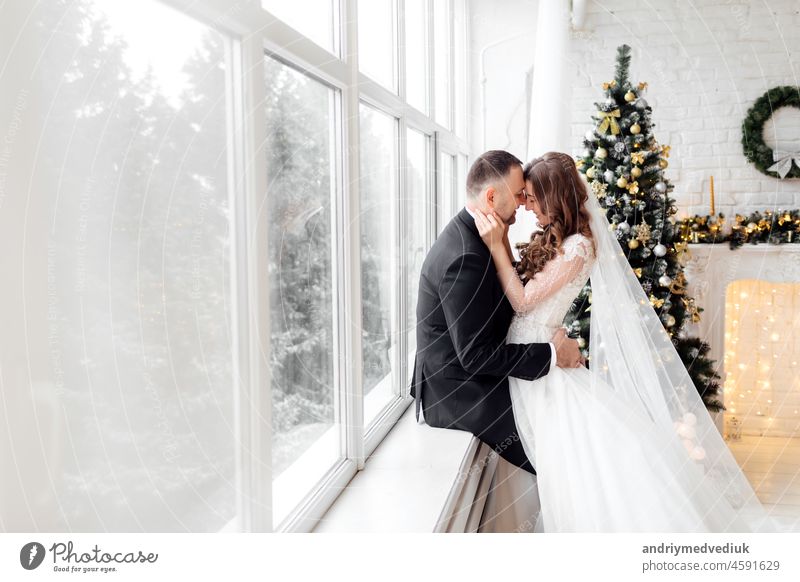 Junges Paar in der Liebe Braut und Bräutigam posieren im Studio auf dem Hintergrund mit Weihnachtsbaum in ihrem Hochzeitstag zu Weihnachten in der Nähe des großen Panoramafenster dekoriert.