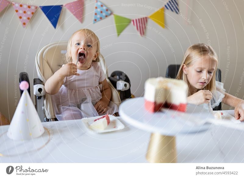 Geburtstagsfeier zu Hause. Glückliche kleine kaukasische Mädchen und niedlichen Baby feiern ersten Geburtstag zu Hause. Candid Lifestyle-Porträt von Kid Blasen Kerzen auf Kuchen. Wohnzimmer mit Fahnen geschmückt.